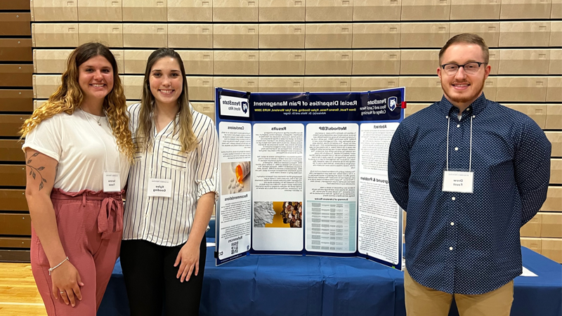 One male and two female students standing by a tri-panel research poster on a table. 